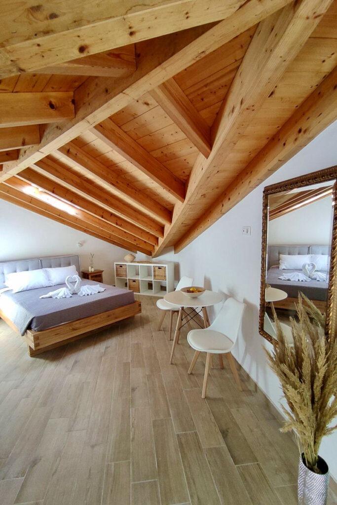 The spacious bedroom of Lysianassa apartment. There's a double bed, a small table with two chairs and a big mirror under the wooden roof.
