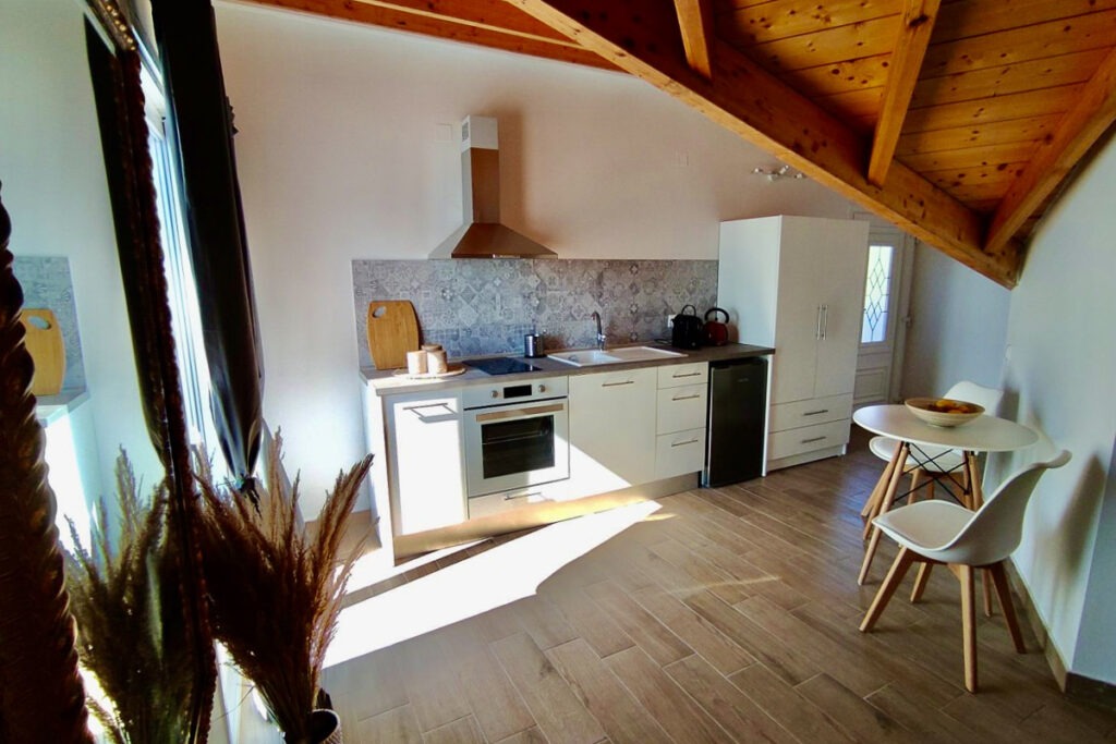 The kitchenette of Lysianassa apartment in the attic with a wooden roof. The kitchenette is white equipped with a cooker, a kitchen hood and a small fridge.