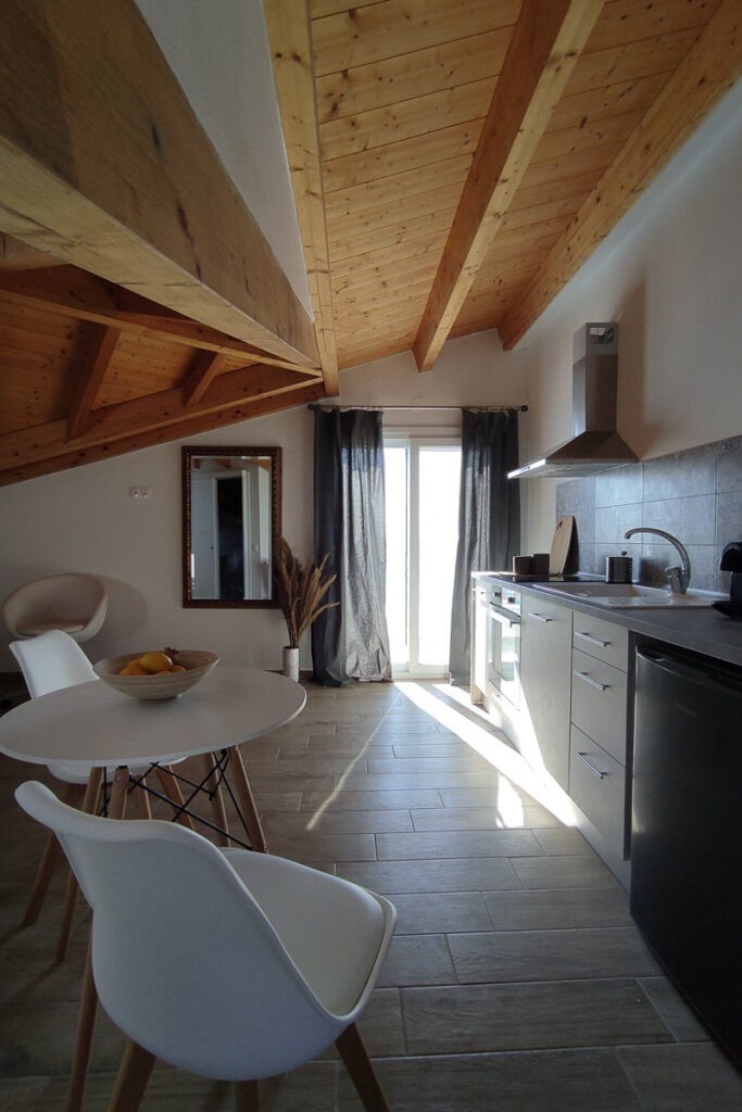 The kitchenette of Lysianassa apartment in the attic with a wooden roof. The kitchenette is white equipped with an oven and a small fridge. There's also a white round table with two white chairs.