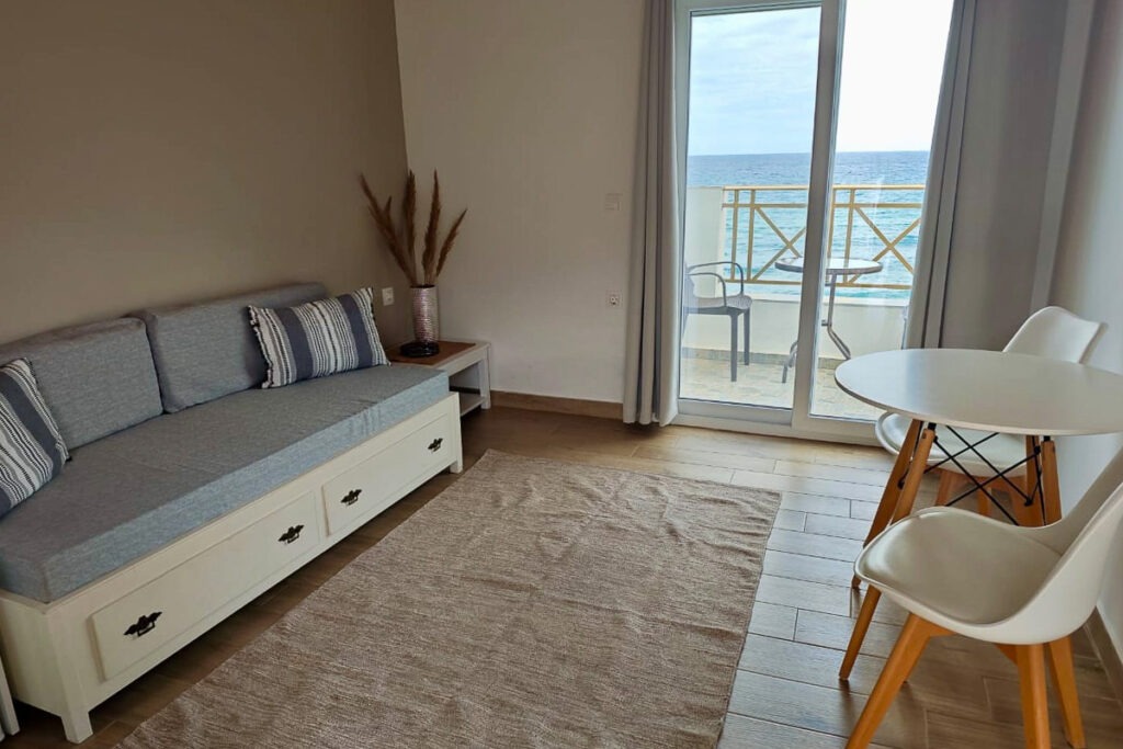 The living room of Nemertes equipped with a white wooden sofa and a white round table with two white chairs. From the living room, there's a sea view.