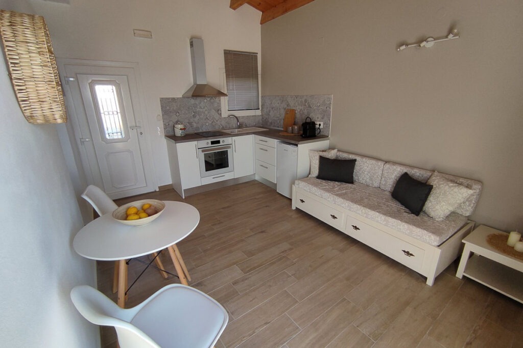 The spacious living room and kitchenette area of Nemertes studio with a white sofa, a white round table with two chairs. The kitchen is white of a corner shape and is equipped with a cooker, a kitchen hood and a small fridge.