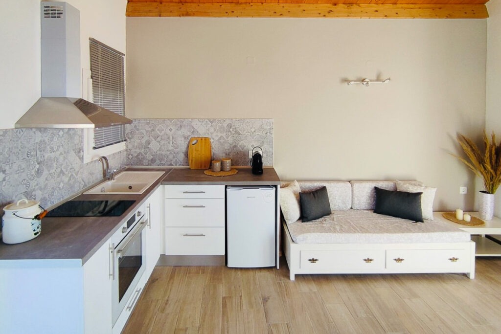The white kitchenette and living room space of Nemertes apartment. A white sofa is next to the kitchen. The kitchen has a corner shape and is equipped with a small fridge, a cooker and a kitchen hood.